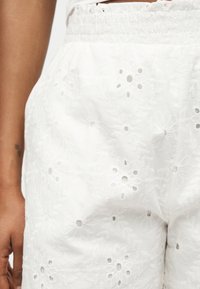 Gros plan d'une personne portant un short blanc taille haute avec une broderie anglaise florale et une ceinture smockée.