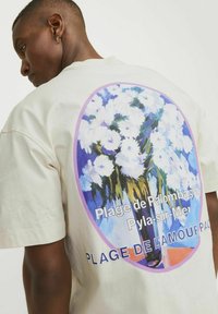 Kreminės spalvos trumpomis rankovėmis t-shirt su mėlynai baltu gėlių grafiniu motyvu ir juodu tekstu. Dizainas yra laisvo ir atsitiktinio stiliaus.