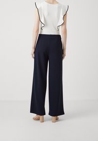 White top with black ruffled trim, paired with high-waisted navy wide-leg pants. Model wearing beige block-heeled sandals.