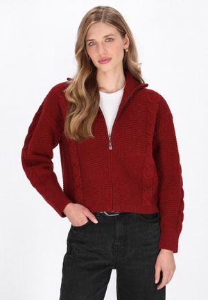 Maglione rosso a zip con un motivo a trecce sulle maniche e sul corpo. Presenta un colletto alto e polsini a costine, abbinato a jeans neri.
