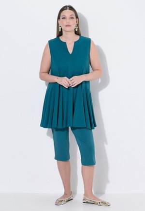 Kobieta stojąca w turkusowej tunice bez rękawów i krótkich legginsach, nosząca kwiatowe baleriny i zielone kolczyki na białym tle.