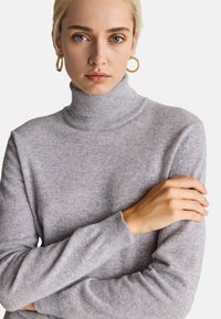 Maglione grigio chiaro a collo alto in morbido tessuto a maglia, con polsini e collo a coste. Il modello posa con le braccia incrociate, mostrando i dettagli delle maniche.