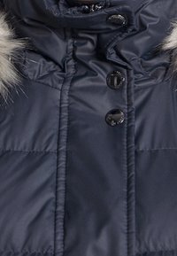 Chaqueta acolchada azul marino con ribete de piel sintética en la capucha, botones negros brillantes, textura acolchada y diseño de cuello alto.