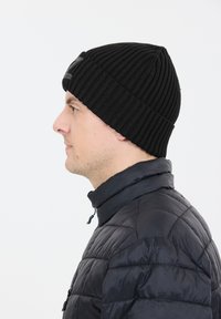 Svart stickad mössa med vertikal ribbning, med en vikbar mudden. Bärs tillsammans med en svart vadderad jacka med hög krage.