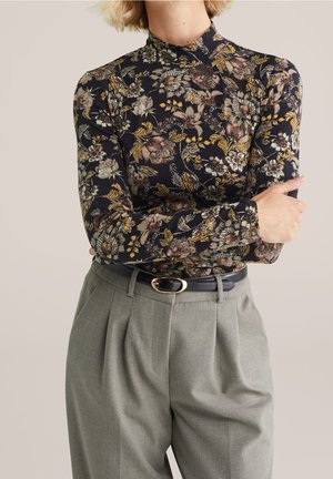 Mujer que lleva una blusa ajustada de manga larga negra con estampado floral, metida dentro de unos pantalones grises de cintura alta con pliegues y un cinturón negro.
