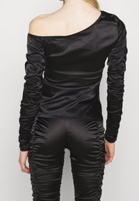 Frau trägt ein gerafftes schwarzes Satin-Top mit schulterfreiem Schnitt und passende geraffte Satinhosen, von hinten vor einem schlichten Hintergrund gezeigt.