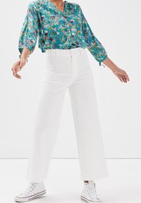 Blouse florale en turquoise avec des manches trois-quarts et des poignets noués, assortie à un pantalon large taille haute blanc et des baskets blanches.