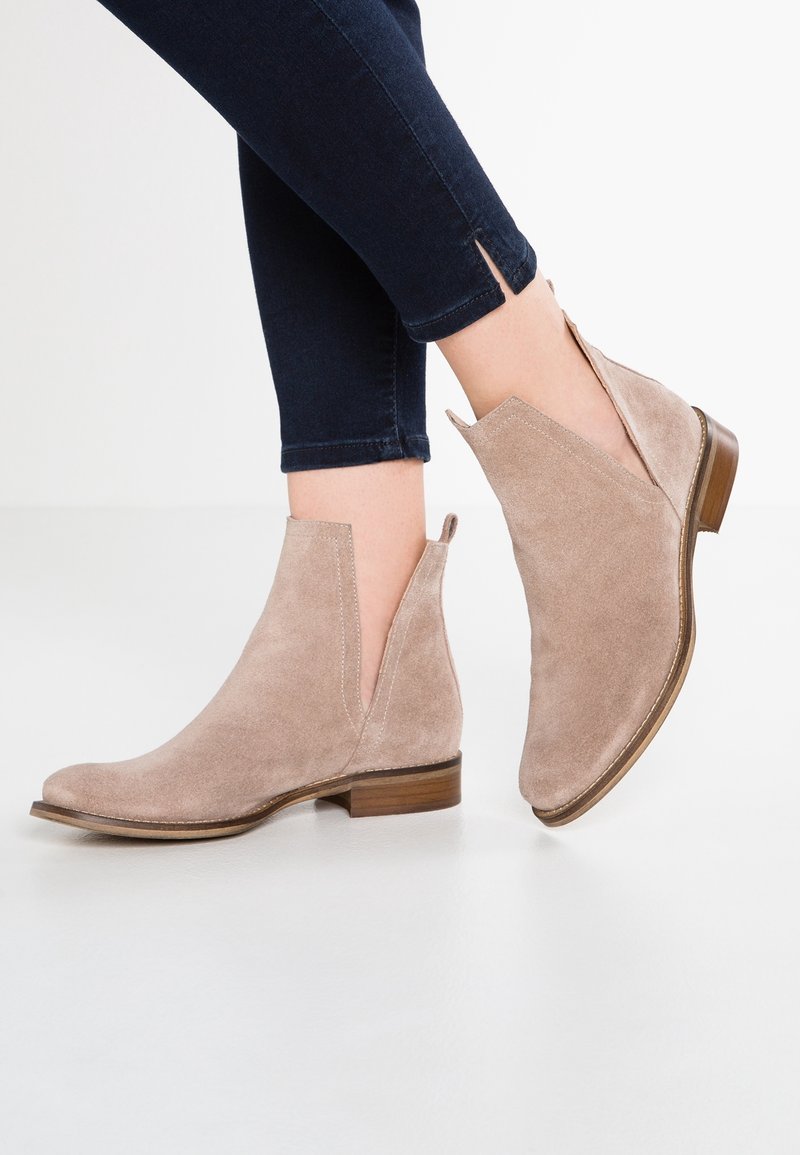 Bottines beiges en daim avec un bout légèrement pointu, des panneaux élastiques sur les côtés et un petit talon empilé, associées à un jean foncé avec des fentes.