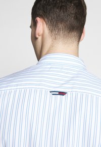 Camicia con bottoni a righe blu chiaro e bianche. Presenta un colletto classico e un piccolo logo ricamato in rosso, bianco e blu sul retro.