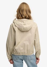 Mujer con cabello rubio hasta los hombros, vistiendo una chaqueta con capucha de color beige claro y jeans azules, vista de espaldas sobre un fondo blanco.