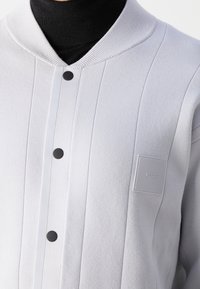 Gros plan d'une veste blanche texturée avec coutures verticales, boutons-pression noirs, col côtelé et écusson rectangulaire avec logo "BOSS" sur la poitrine.
