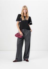 Femme debout dans un pantalon gris large, un haut noir avec des bordures blanches, tenant un sac à main bordeaux et portant des chaussures bordeaux assorties.
