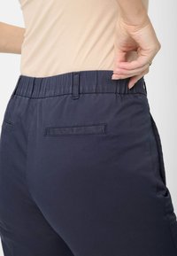 Marineblaue Hose mit elastischem Bund und glatter Textur sowie hinteren Taschen, seitlich gezeigt mit einem teilweise sichtbaren hellbeigen Oberteil.