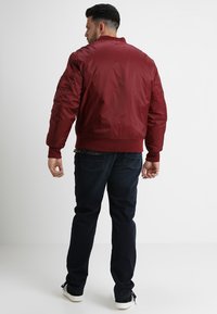 Burgunderfarget bomberjakke med en glatt, shiny tekstur, ribbede ermer og linning. Kombinert med mørkeblå jeans og enkle hvite joggesko.