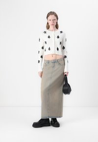 Donna che indossa un cardigan corto in maglia bianca con motivi floreali neri, una lunga gonna di denim beige, scarpe platform nere e tiene una borsa nera.