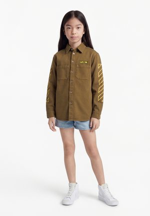 Young person standing wearing a brown button-up shirt with yellow sleeve designs, denim shorts, and white high-top sneakers.