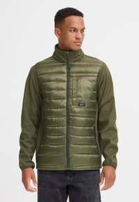Jachetă verde căptușită cu guler în picioare, având un fermoar frontal și o buzunar pe piept. Materialul este nylon cu o textură netedă.