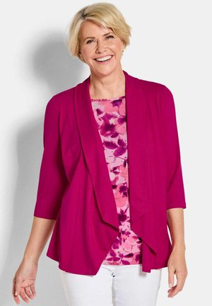 Lächelnde Frau mittleren Alters, die eine leuchtend pinke, offene Strickjacke über einem floralen pinken Oberteil und weißen Hosen trägt, vor einem einfachen Hintergrund stehend.