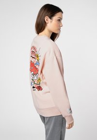 Hellrosa Sweatshirt mit rundem Ausschnitt, auf der Rückseite mit bunten Cartoon-Grafiken versehen, mit gerippten Bündchen und einem Etikett am Ärmel.