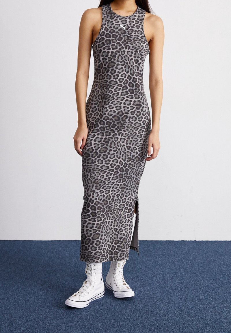 Femme portant une robe maxi grise sans manches à imprimé léopard avec une fente latérale, associée à des baskets blanches montantes sur un tapis bleu.