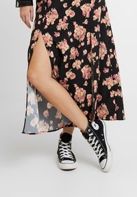 Jupe longue noire à fleurs avec fente latérale, ornée de fleurs roses et crème. Associée à des baskets montantes Converse noires.