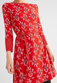 Punainen pitkähihaista mekkoa, jossa on kukkakuosi ja lehtikoristeita. Materiaali on pehmeää, vyötäröllä solmittava nauha ja hihoissa on pilkkukuvioita.