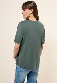 T-shirt verde in cotone con maniche corte arrotolate, collo a giro e vestibilità rilassata. Presenta una texture liscia e un orlo leggermente curvato.