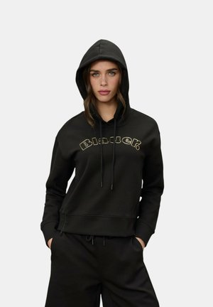 Donna che indossa una felpa nera con il cappuccio alzato e il testo "Blauer" sul petto, abbinata a pantaloni neri e mani nelle tasche.
