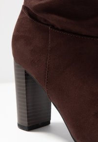 Brun mockaskenkelstövel med en fyrkantig klack med randig design. Syt detaljer synliga längs den övre sömmen.