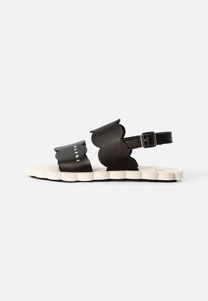 Sandálias de couro pretas com correias em forma de ondas e uma fivela, apresentando um sola de borracha branca texturizada com um design de borda ondulada.