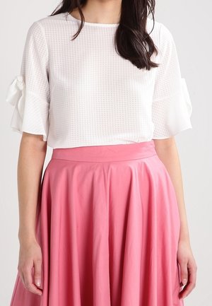 Kobieta ubrana w białą, teksturowaną bluzkę z krótkim rękawem z kokardkami oraz wysoką, różową spódnicę o luźnym kroju.