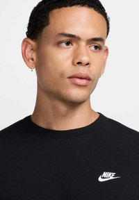 Felpa nera a girocollo in cotone con texture a coste. Presenta un logo Nike bianco sul petto. Il modello ha i capelli corti e ricci.