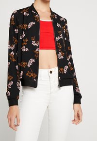 Svart zip-up bomberjacka med rosa och bruna blommönster över en röd kort topp och vita högmidjade jeans på stående person.