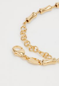 Gouden kettingarmband met cilindrische schakels, ronde kettingverlenging, musketonhaak en gegraveerde "GUESS" logo plaat op witte achtergrond.