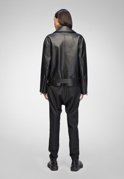 Schwarze Lederjacke mit breiten Revers; kombiniert mit schwarzen, locker sitzenden Hosen. Die Jacke hat eine taillierte Form mit Gürtel und Reißverschlüssen an den Ärmelabschlüssen.