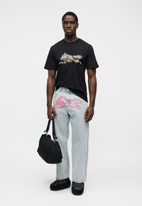 Μαύρο πουκάμισο με κοντά μανίκια και γραφικό σχέδιο, ανοιχτό μπλε φαρδιά τζιν με ροζ καρτούν γάτα, μαύρη τσάντα και σκούρα υποδήματα.