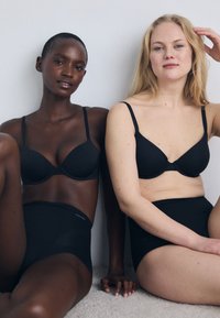 Black bras with adjustable straps, featuring a smooth texture and a basic design; worn with high-waisted black underwear. Two models pose together.