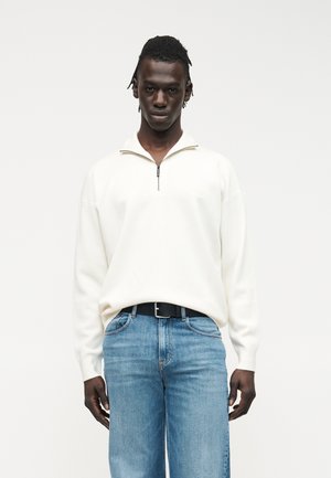 Černoch stojí, oblečený v bielom polozipsom svetri a svetlomodrých džínsoch s čiernym opaskom, na jednoduchom bielom pozadí.