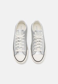 Zapatillas de lona plateadas con punta de goma blanca, franja de acento negra y cordones crema. Presenta el logo "Converse All Star" en la lengüeta.