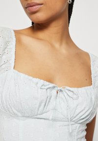 Blusa blu chiaro ricamata con motivi floreali, colletto quadrato, maniche a sbuffo e dettaglio con fiocco frontale. Tessuto morbido con un motivo floreale testurizzato.