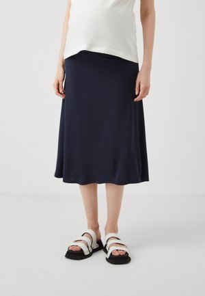 Gonna midi blu navy realizzata in morbido tessuto drappeggiato con una superficie liscia; abbinata a sandali strappy bianchi e neri con suola spessa.