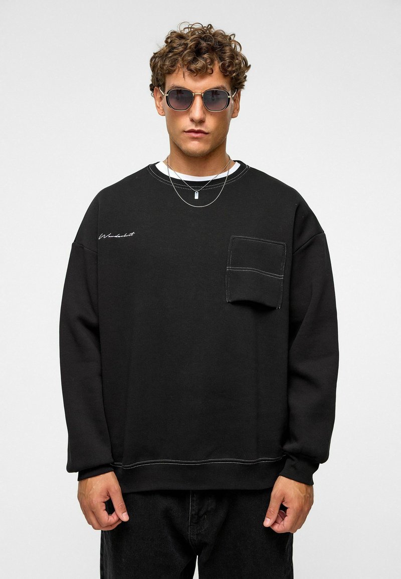 Zwart sweatshirt met een ronde halslijn, een voorzak en witte stiksels. Draagt over een wit T-shirt en gecombineerd met een zonnebril.