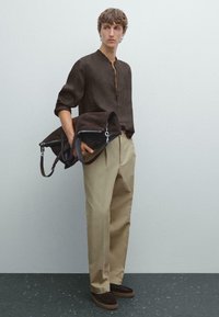 Brown suede bag with silver hardware held by a person wearing a dark brown shirt and beige trousers, standing on a textured gray surface.