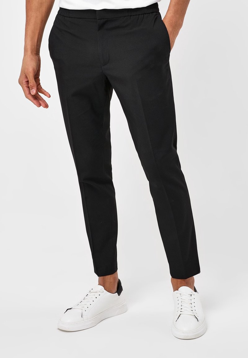 Homme portant un pantalon noir slim et des baskets blanches, debout les mains dans les poches sur fond blanc.