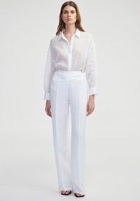 Camicia bianca con bottoni e maniche lunghe, abbinata a pantaloni bianchi a vita alta con dettagli in metallo argentato e taglio su misura.