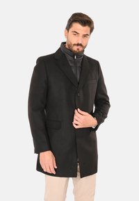 Uomo con barba che indossa un cappotto nero con bottoni sopra una giacca trapuntata nera e pantaloni beige, che si sistema il cappotto con una mano.