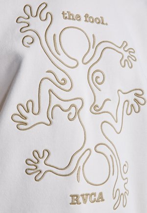 Fehér szövet dombornyomott aranyszállal, amely egy karokkal és lábakkal rendelkező alakot ábrázol. Fölötte a "the fool." felirat olvasható, alatta pedig az "RVCA".