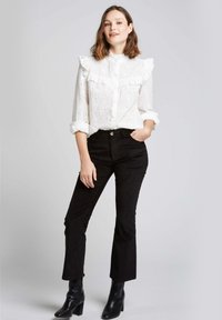 Blouse blanche avec un col à volants et des détails en œillets, associée à un pantalon en velours côtelé noir slim-fit et des bottines noires.