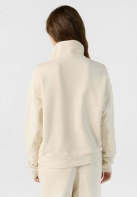 Lichtbeige pullover met een hoge kraag en lange mouwen. Gemaakt van zacht stof, heeft het een ontspannen pasvorm en ribbels aan de manchetten.