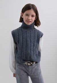Pige med skulderlangt brunt hår iført en grå ærmeløs turtleneck sweater, hvid langærmet skjorte, grå bukser og et mønstret bælte.